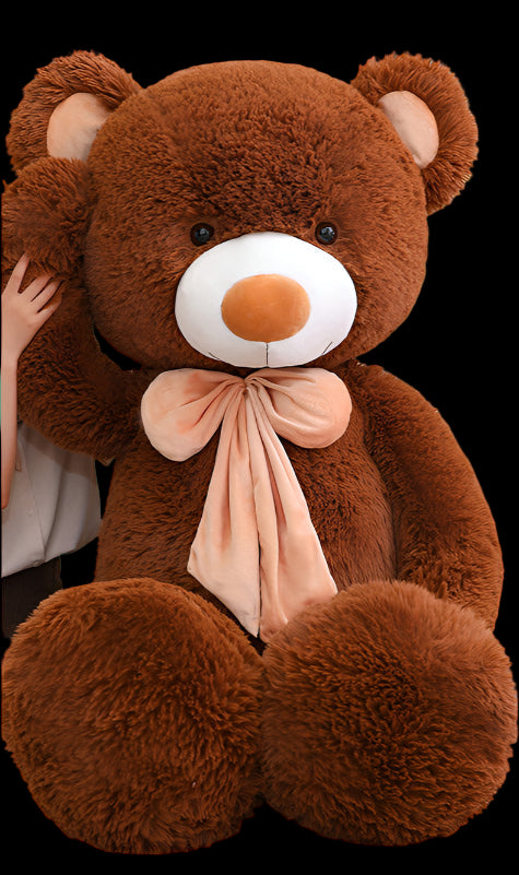 Person sitting next to a large brown teddy bear with a pink bow in a room.