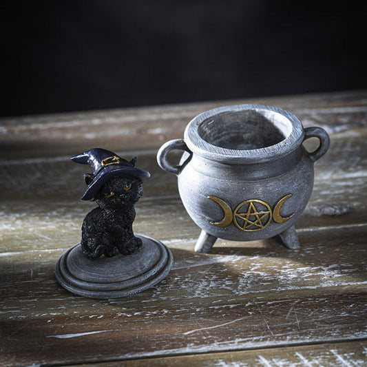 Black cat figurine wearing a witch hat next to a cauldron with wiccan symbols on a wooden surface.