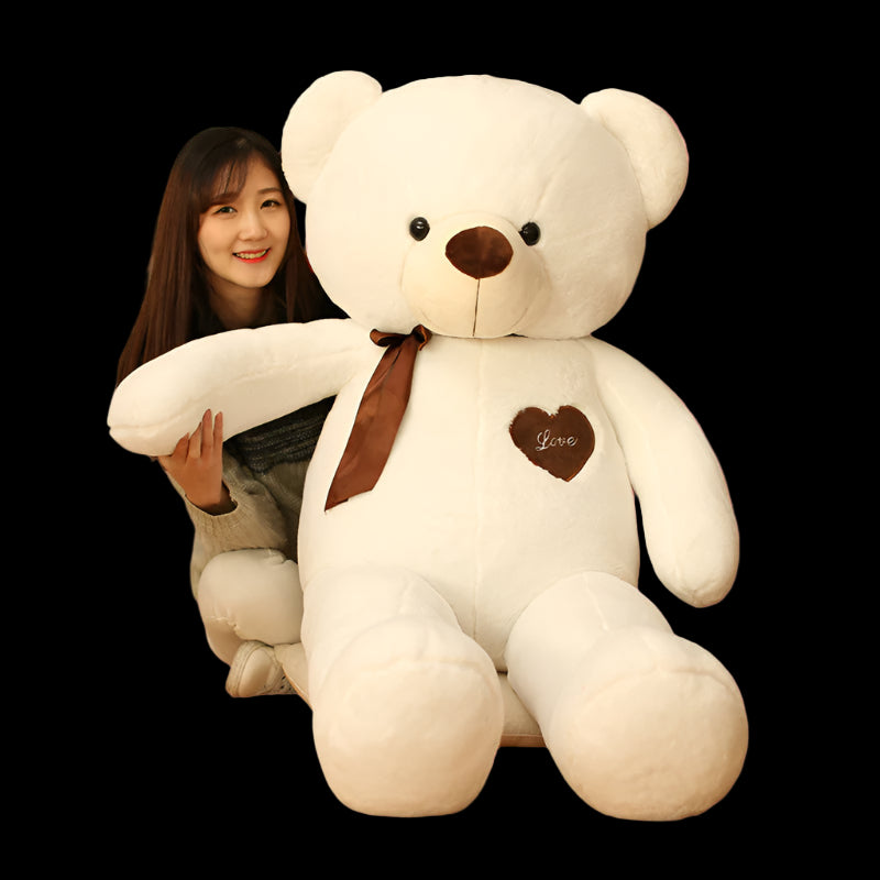 Woman holding a large white teddy bear with a brown bow tie and heart emblem indoors.