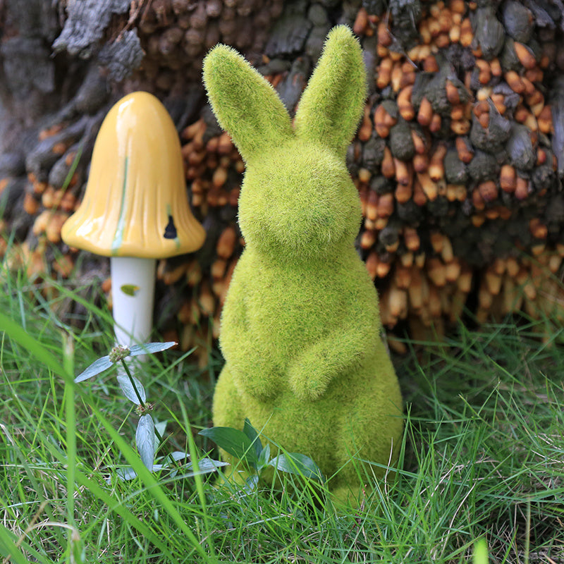 Green mossy rabbit figurine and yellow mushroom in a natural setting with grass and woodpile.