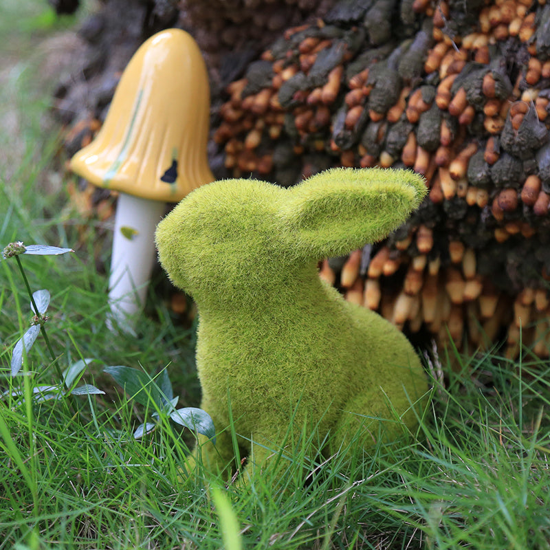 Moss-covered rabbit figurine in a natural setting with a yellow mushroom and pinecones.