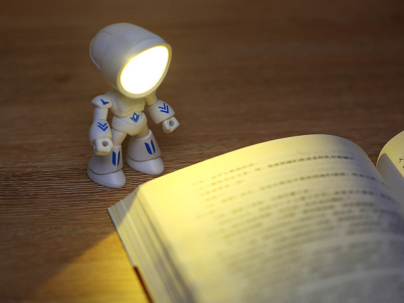 Toy robot with a light on its head illuminating an open book on a wooden surface