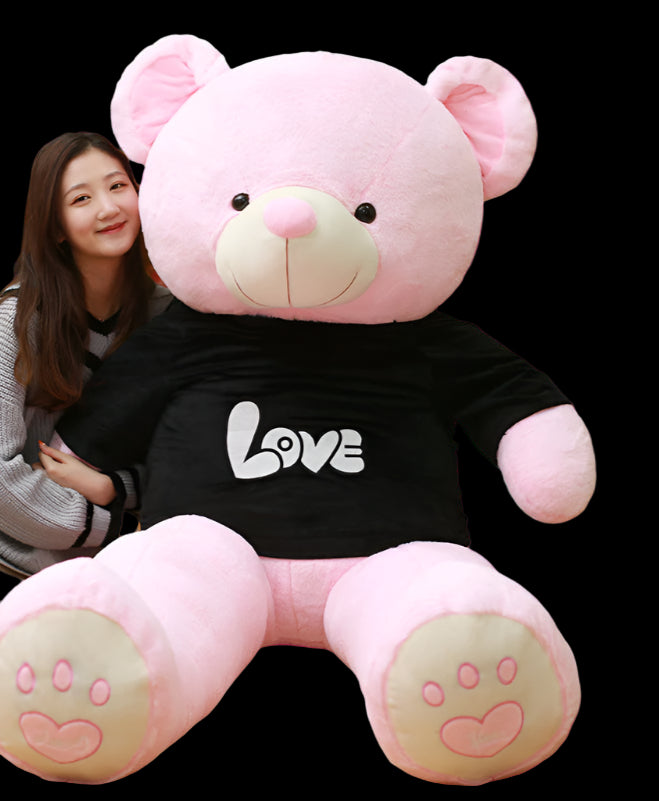 Person holding a large pink teddy bear with 'LOVE' on a black shirt in front of a bookshelf.
