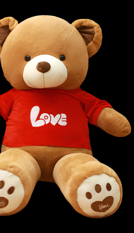 Woman sitting next to a large teddy bear wearing a red 'LOVE' shirt in a room with bookshelves.