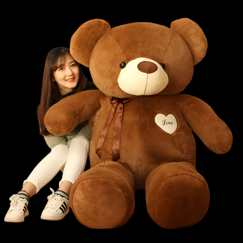 Large brown teddy bear with a heart and ribbon, sitting on a wooden floor with a person next to it.