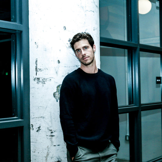 Man in a black sweater leaning against a textured wall with large windows.