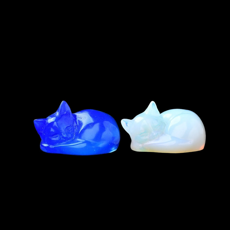 Two cat-shaped figurines, one blue and one white, on a dark surface with a blurred green plant in the background.