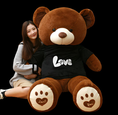 Large brown teddy bear wearing a black shirt with 'LOVE' on it, sitting next to a person in a library setting.