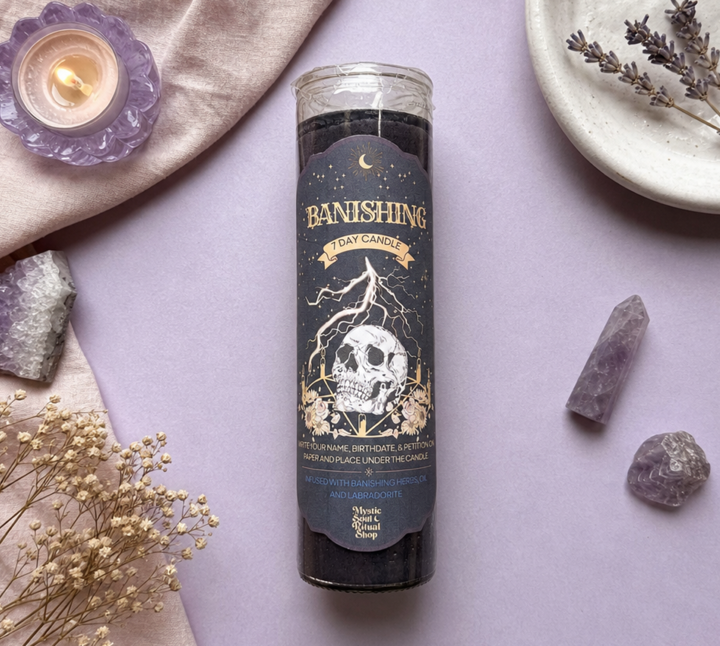 A black tarot card candle in a clear glass jar labeled 'Banishing Day Candle' by Magic Shop, styled in a flat lay on a lavender background with crystals, dried flowers, and a small lit candle.