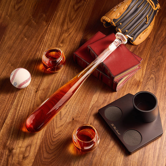 Bar spoon, baseball, glass of amber liquid, baseball glove, and book on a wooden surface