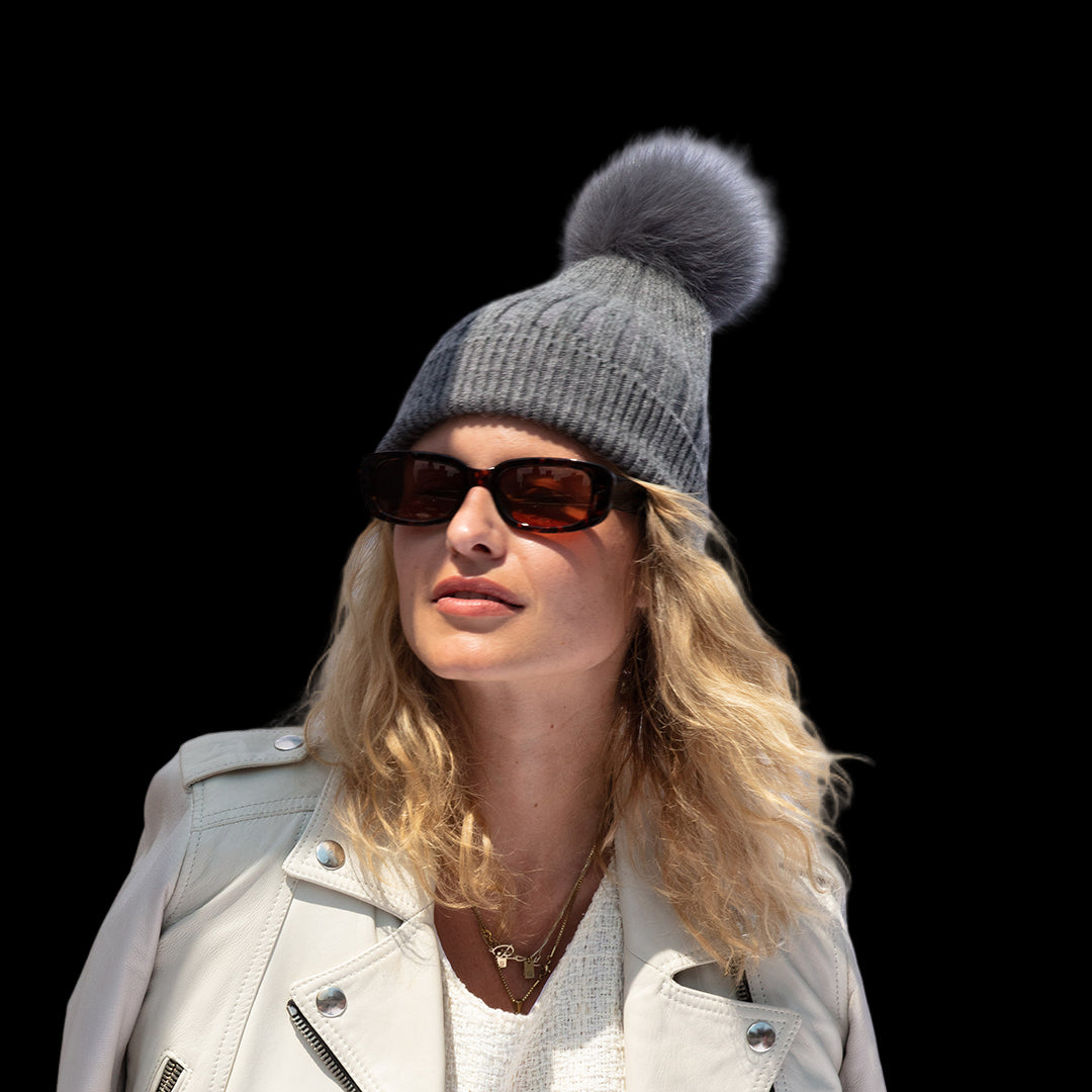 Woman wearing a gray knit beanie with a pom-pom, sunglasses, and a light-colored jacket against a white curtain background.