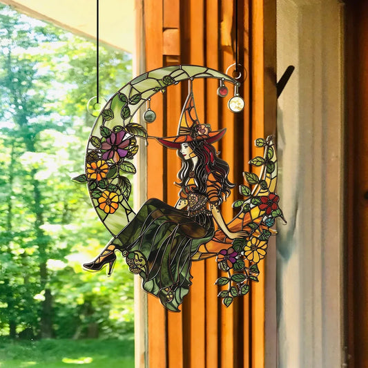 Decorative stained glass panel with a witch sitting on a moon, surrounded by flowers, hanging in front of a window with a view of trees.