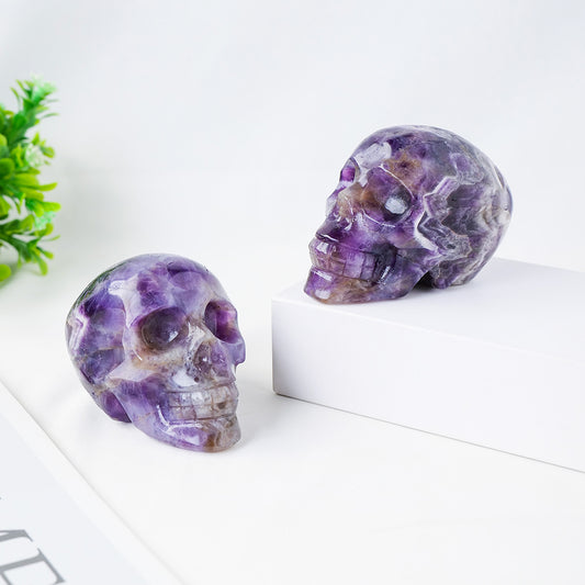 Two purple crystal skulls on a white surface with a green plant in the background.
