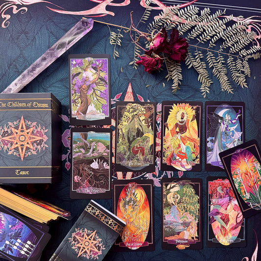 A styled flat-lay of The Children of Osiris Tarot deck, showing the main box, several cards laid out, a crystal ball, dried roses, and greenery on a dark patterned fabric background.