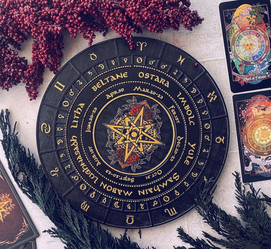 A circular black and gold Celtic calendar wheel with zodiac and moon phases, styled in a flat lay with red berries, pine branches, and tarot cards on a light stone surface.