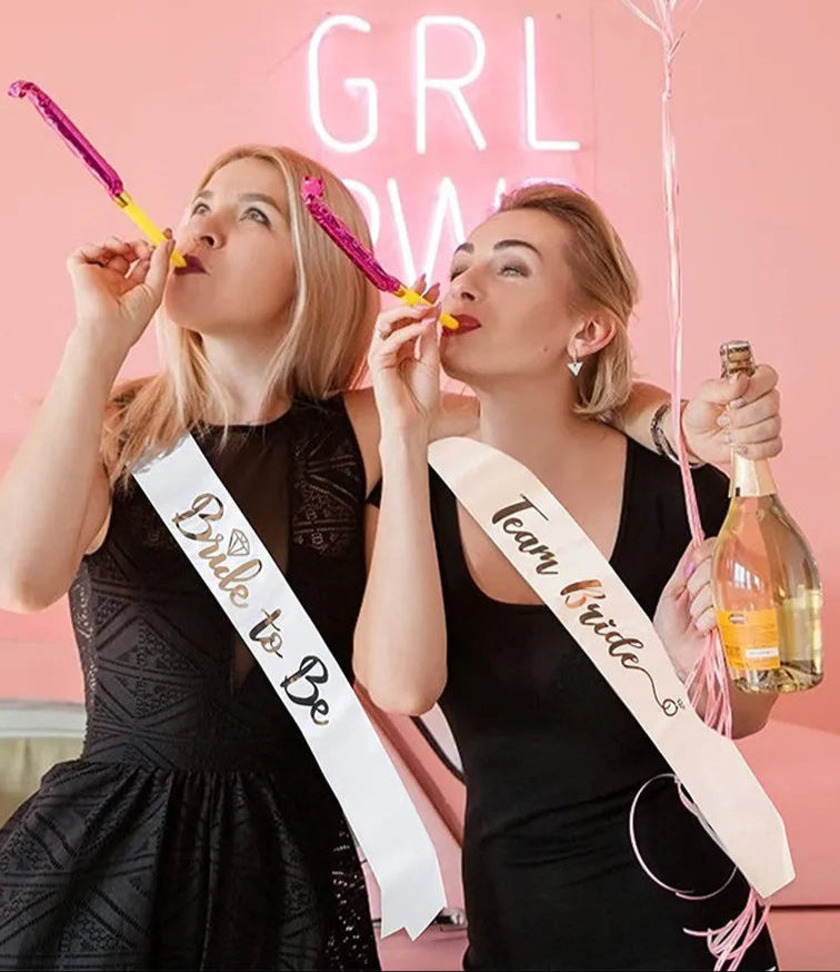 Two women celebrating with 'Bride to Be' sashes against a pink background with 'GIRL POWER' neon sign.