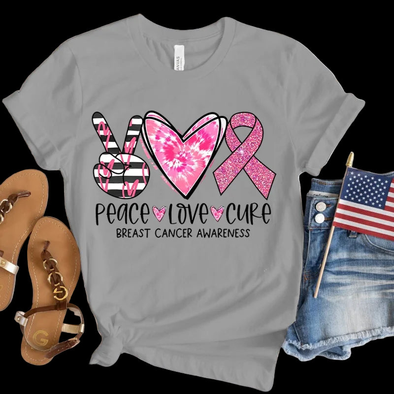 Gray t-shirt with peace, love, cure breast cancer awareness design on a wooden surface with sandals and an American flag.