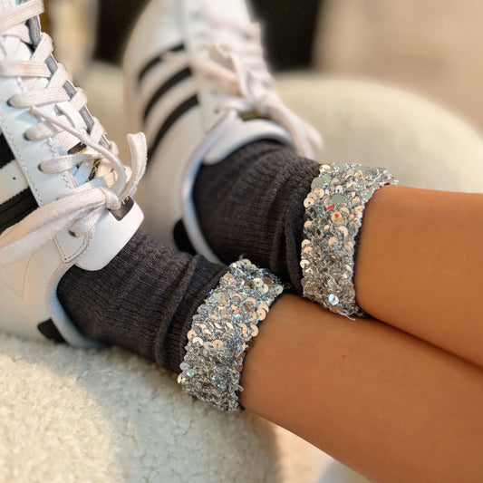 Close-up of white sneakers with black stripes and brown socks, worn by a person with glittery silver bracelets.