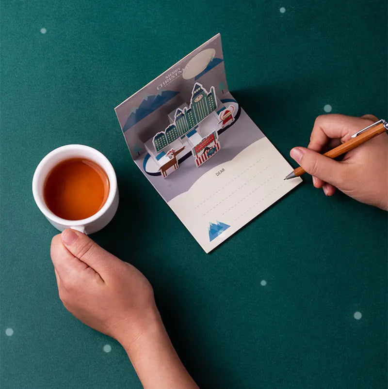 Person holding a cup of tea and writing on a pop-up card with a scenic design.