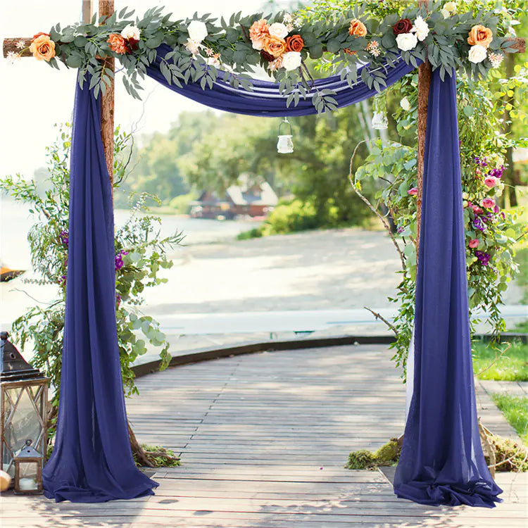 Decorative arch with flowers and purple curtains on a wooden deck by a lake.