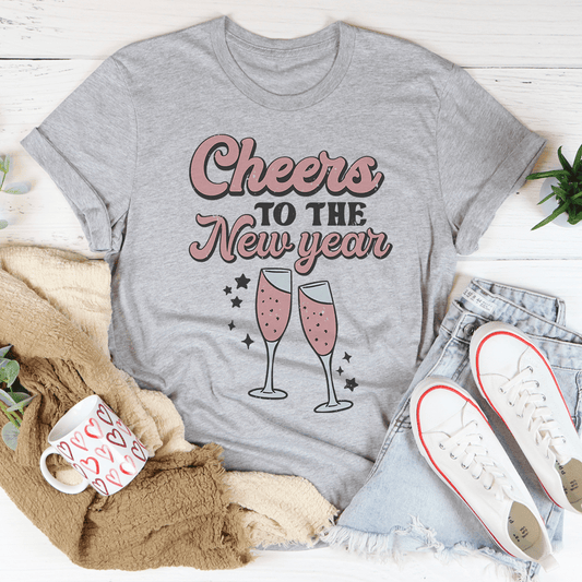 Gray t-shirt with 'Cheers to the New Year' text and champagne glass graphics, surrounded by casual items on a light wooden surface.