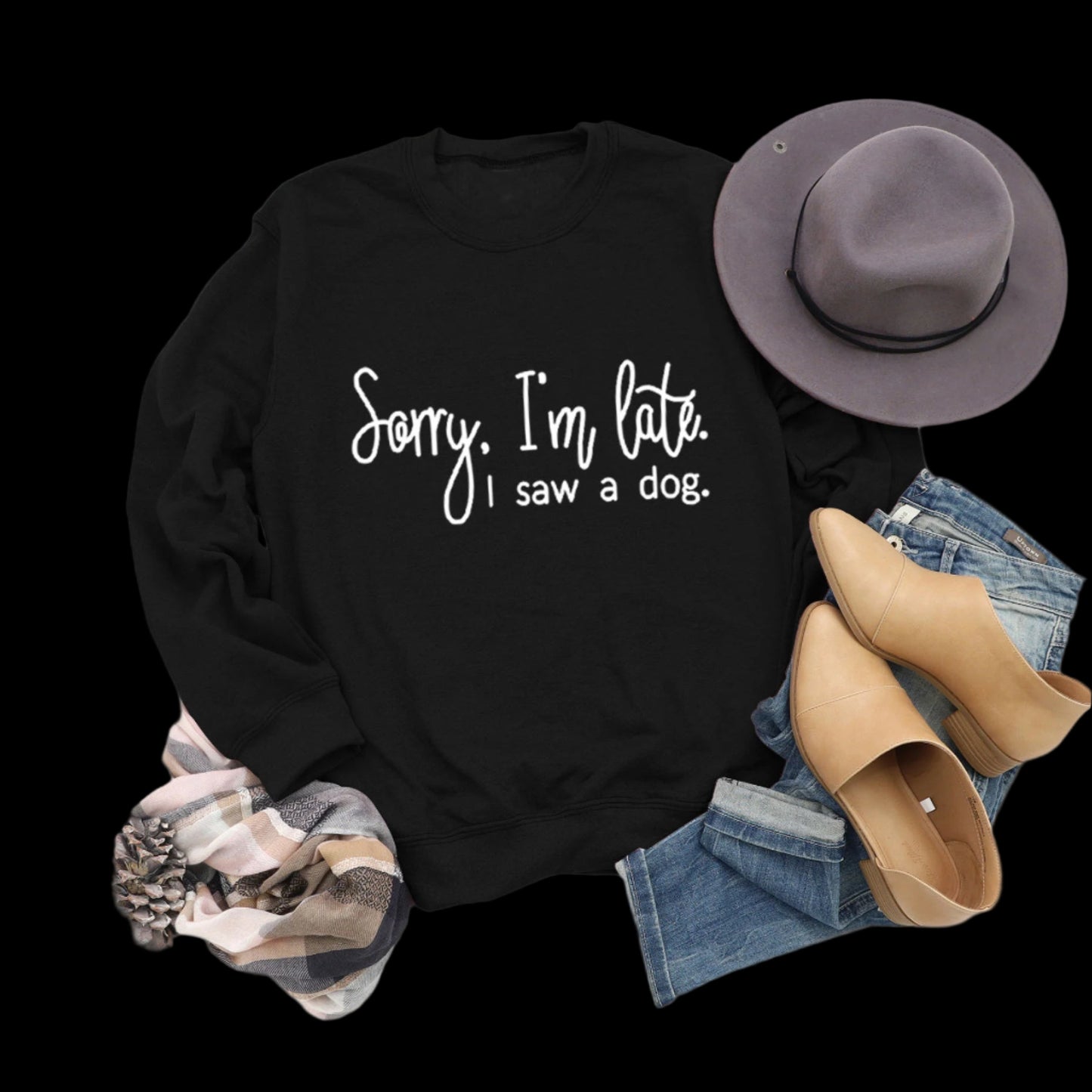 Black sweatshirt with 'Sorry, I'm late. I saw a dog.' text, paired with jeans, boots, and a hat on a wooden surface.