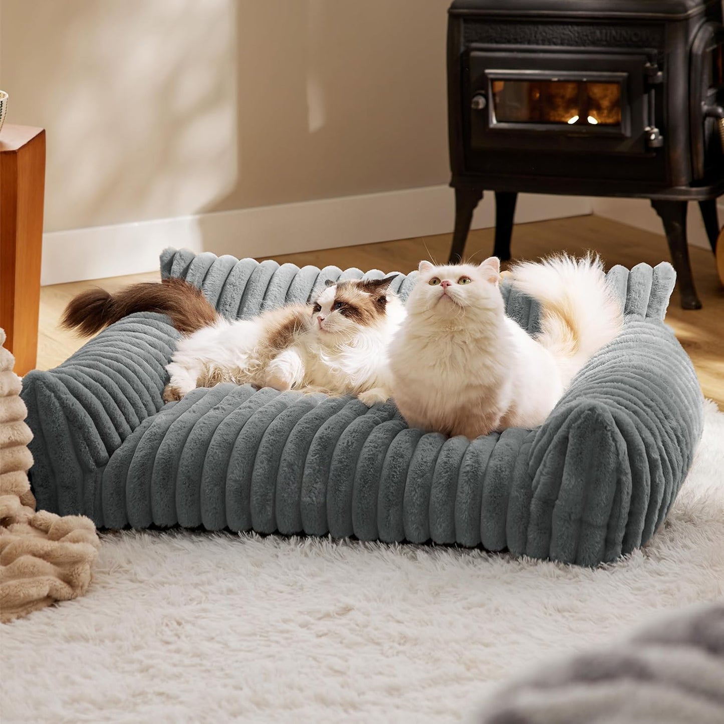 Two cats lounging on a gray pet bed in a cozy room with a fireplace.
