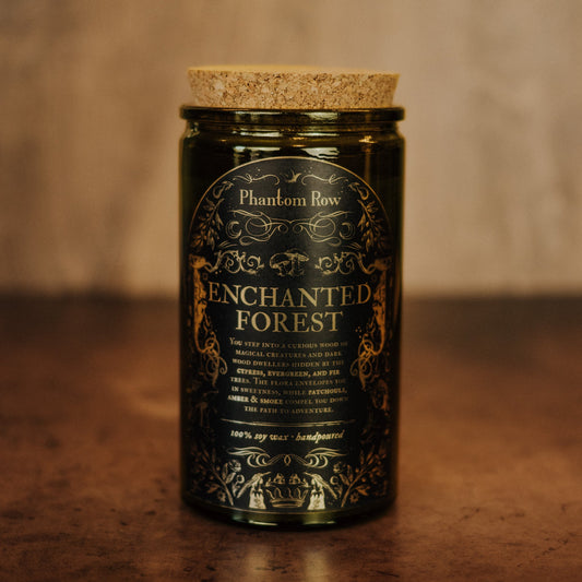 A dark amber glass jar candle with a cork lid and a black label featuring gold ornate detailing that reads 'Phantom Row Enchanted Forest' on a wooden surface.