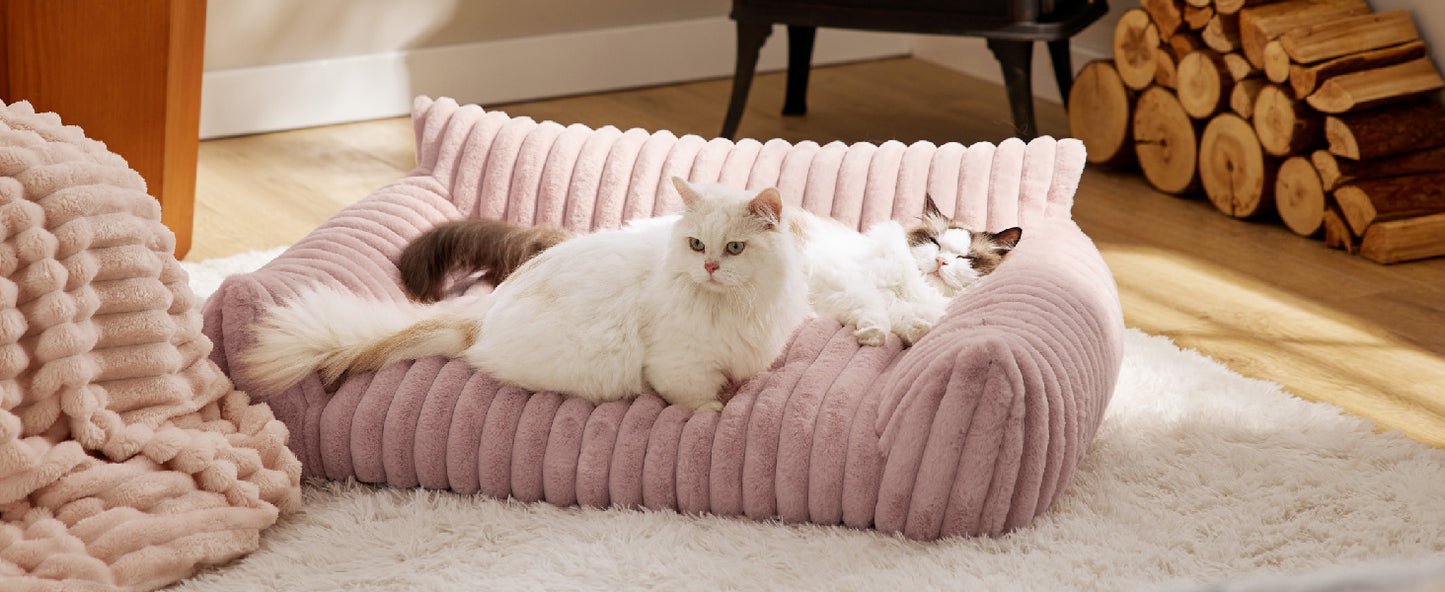 Three cats on a pink ribbed pet bed in a cozy room with wooden logs and a chair.