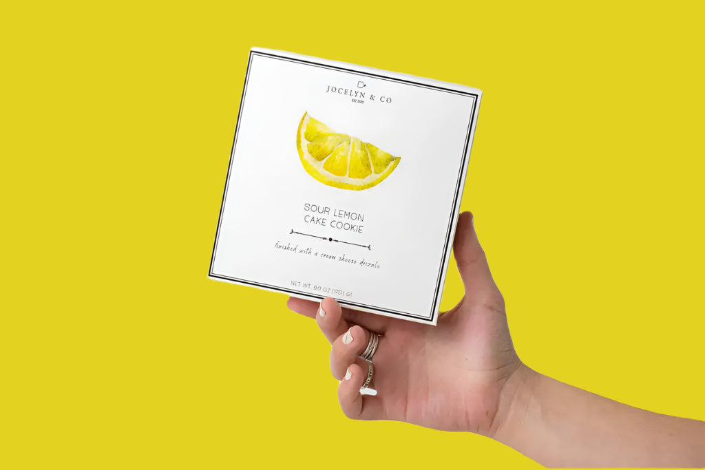 Hand holding a box of sour lemon lime cookies with a lemon illustration on a white background