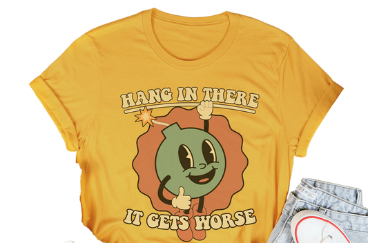 Yellow t-shirt with cartoon character and text 'Hang in there, it gets worse' on a white surface with casual items.