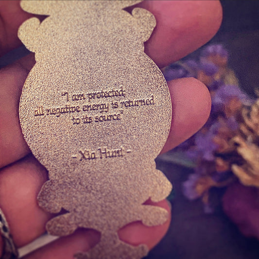 A close-up of a hand holding a glittery gold metal charm shaped like a cartoon character. The charm features an engraved quote by Xia Huji.