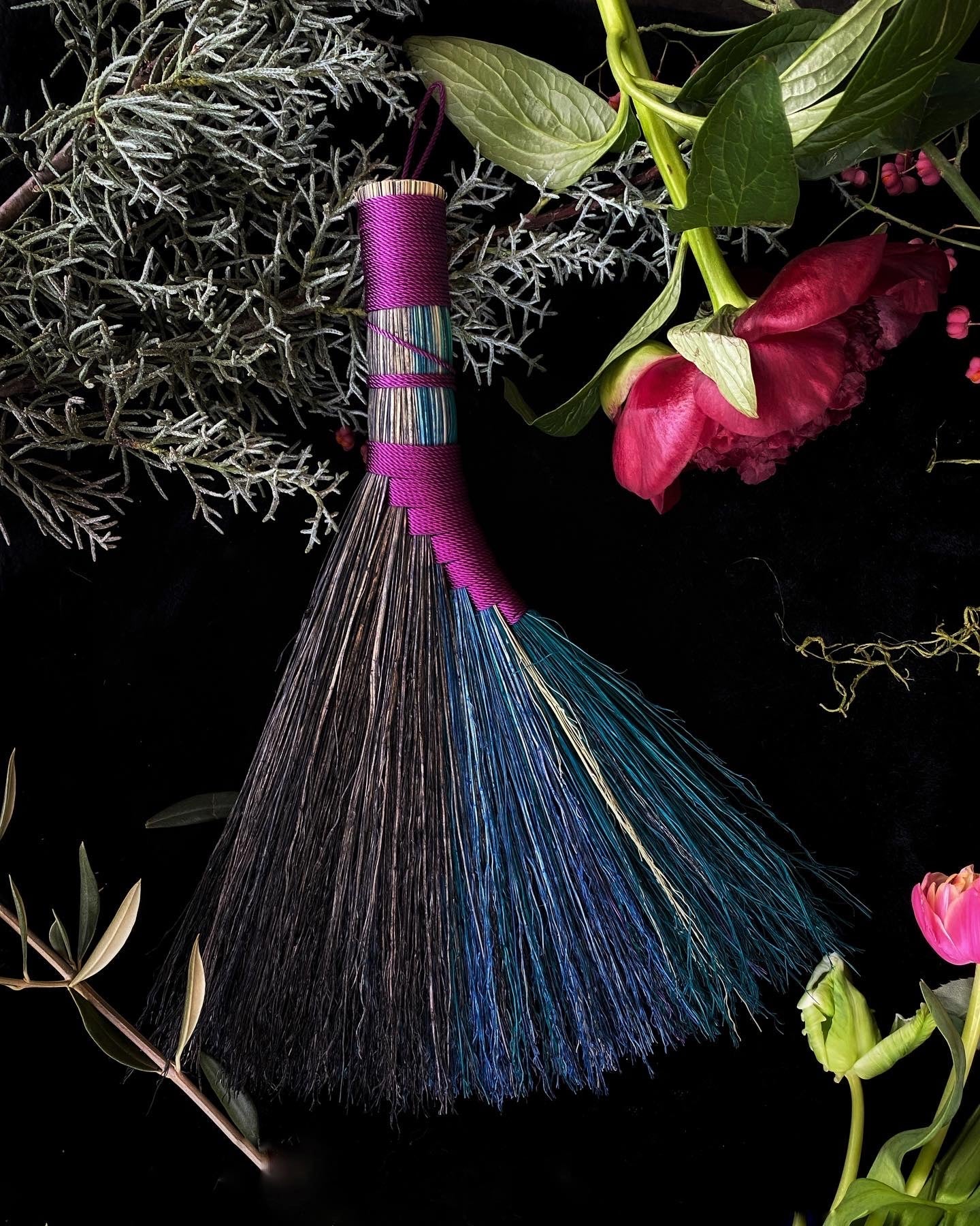 Broom with pink handle and blue bristles on a dark background with greenery