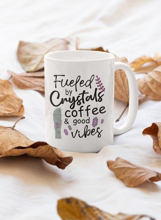 White mug with text 'Fueled by crystals, coffee & good vibes' on a white background with dried leaves.