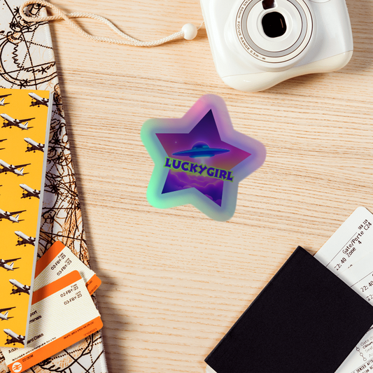 Holographic star-shaped sticker with 'LUCKYGIRL' text on a wooden surface with a camera and travel items.