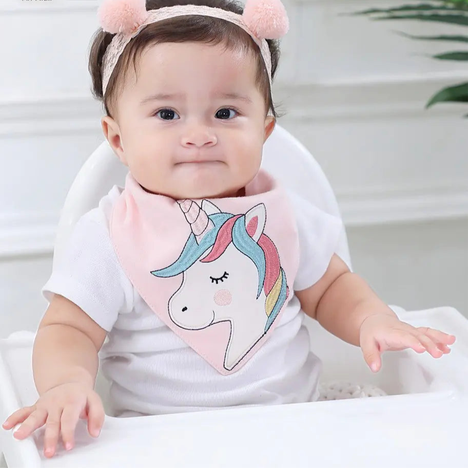 Baby wearing a unicorn-themed bib and headband with pink pom-poms, sitting on a white surface.