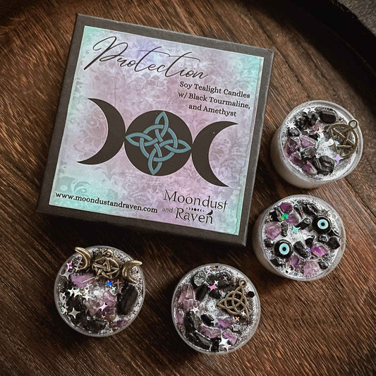 A boxed set of Moondust and Raven Protection soy twilight candles with black tourmaline and amethyst, shown with three small open candles on a dark wood surface. The box features a pastel moon-phase and triquetra design, and the candles contain silver glitter, black stones, purple crystals, and small metal charms.