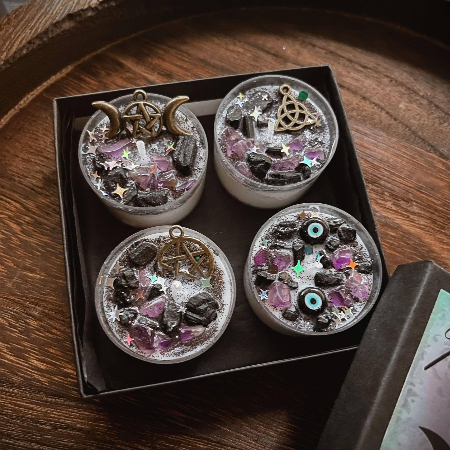 Four small white candles arranged in a square black gift box, each topped with silver glitter, black stones, pink crystal pieces, and a different metallic occult symbol charm including a triple moon, pentagram, knotwork, and eye of providence.
