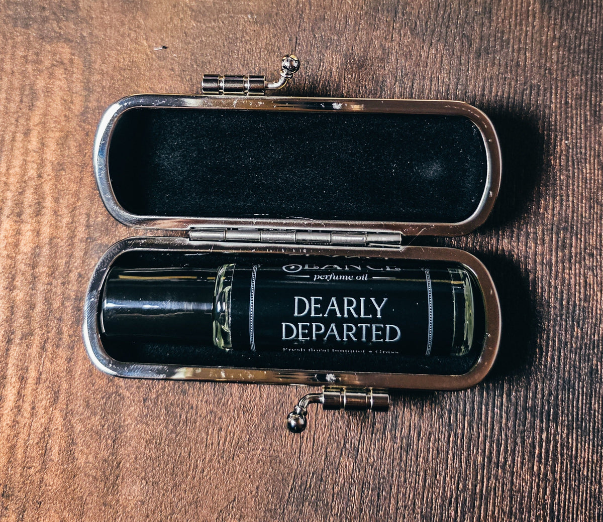 Open case with a perfume bottle labeled 'Dearly Departed' on a wooden surface
