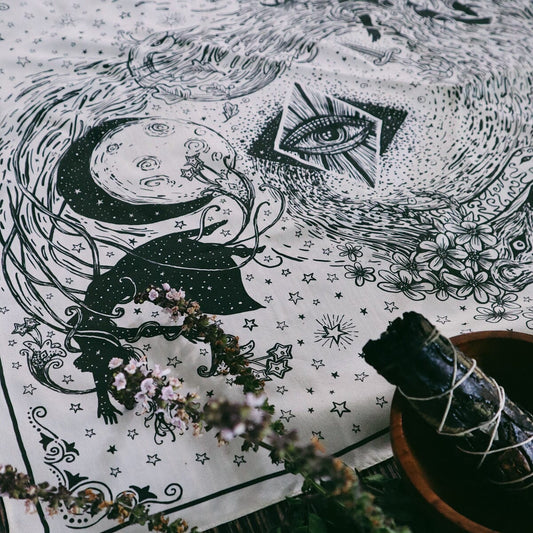 Black and white intricate design with mystical symbols on a textured surface, accompanied by plants and a sage bundle.