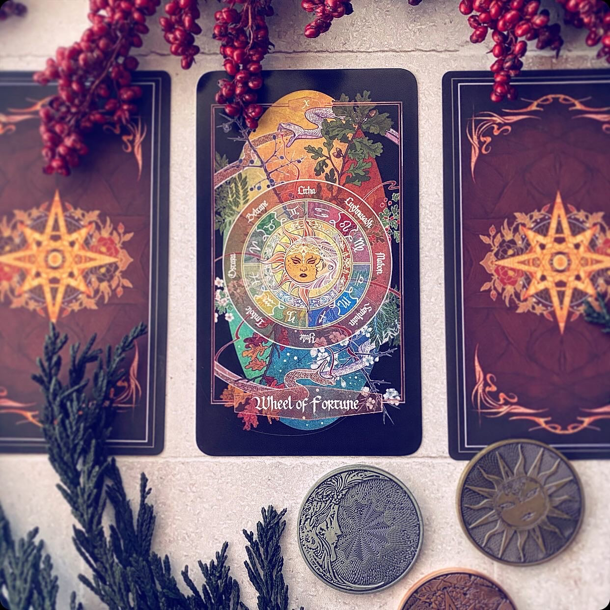 A tarot card deck displayed on a light stone surface, featuring the 'Wheel of Fortune' card in the center with two matching cards behind it. The cards have a dark reddish-brown background with gold starburst designs.