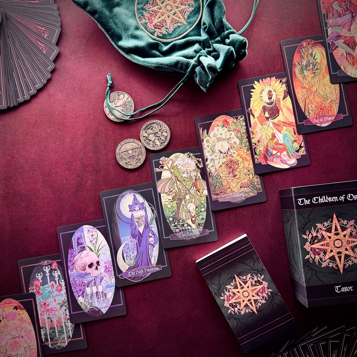 A styled flat-lay of a tarot card deck and accessories on a deep burgundy fabric background. Visible items include multiple illustrated tarot cards, a black box labeled 'The Children of Osiris Tarot', a green velvet drawstring bag, several silver coins, and a partial matching fan.