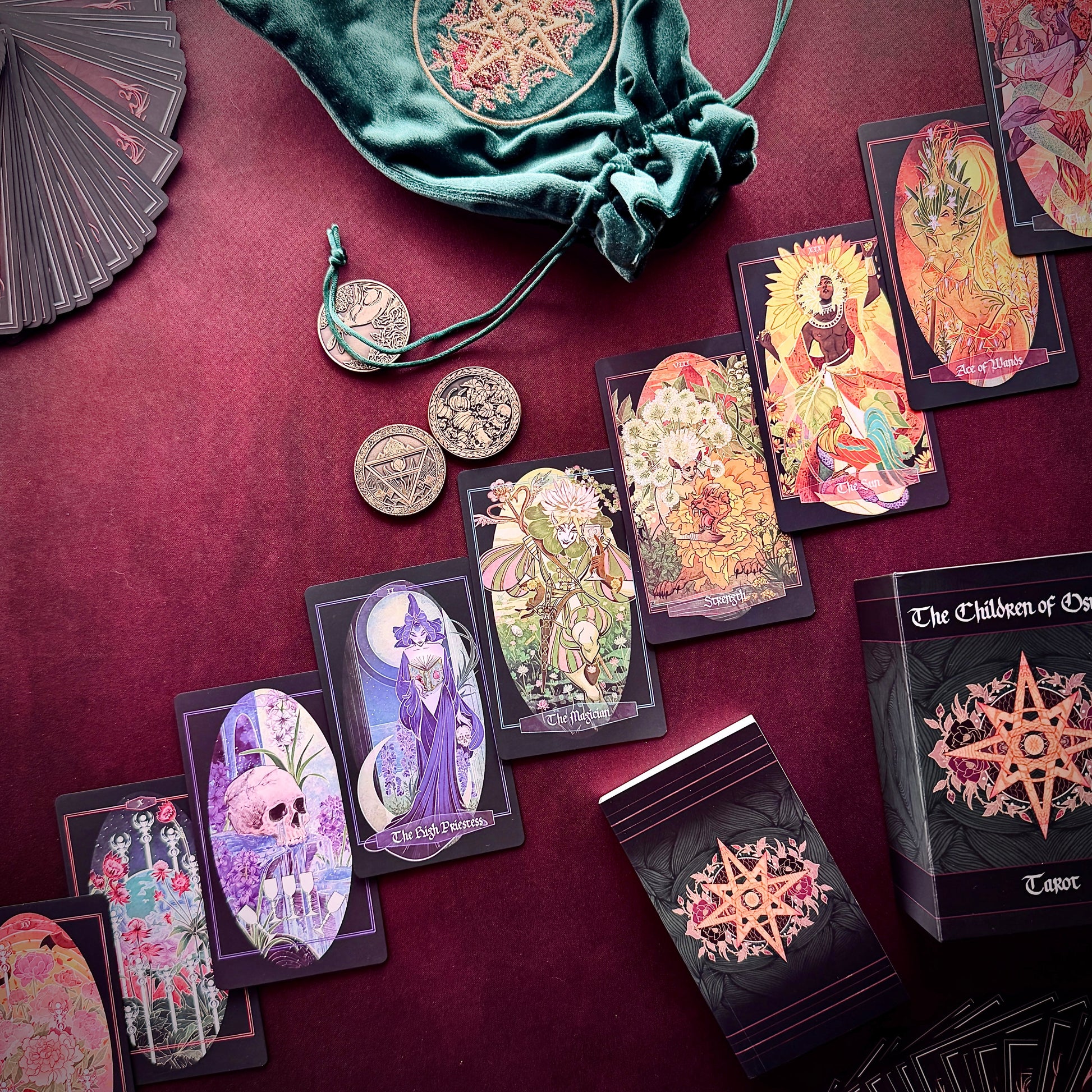 A styled flat-lay of a tarot card deck and accessories on a deep burgundy fabric background. Visible items include multiple illustrated tarot cards, a black box labeled 'The Children of Osiris Tarot', a green velvet drawstring bag, several silver coins, and a partial matching fan.