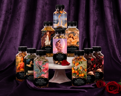 A collection of ten decorative bottles of essential oils and body scrubs arranged on a white marble pedestal against a white background. The bottles feature artistic illustrations of classical figures, fruits, and spices, and are topped with black caps.