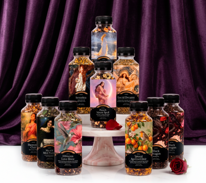 A collection of ten decorative bottles of essential oils and body scrubs arranged on a white marble pedestal against a white background. The bottles feature artistic illustrations of classical figures, fruits, and spices, and are topped with black caps.