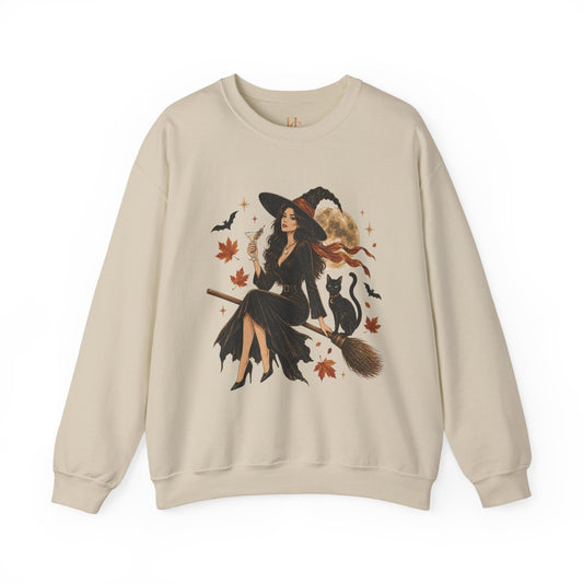 A beige crewneck sweatshirt featuring a large graphic print of a witch riding a broomstick with a black cat, surrounded by autumn leaves and bats.