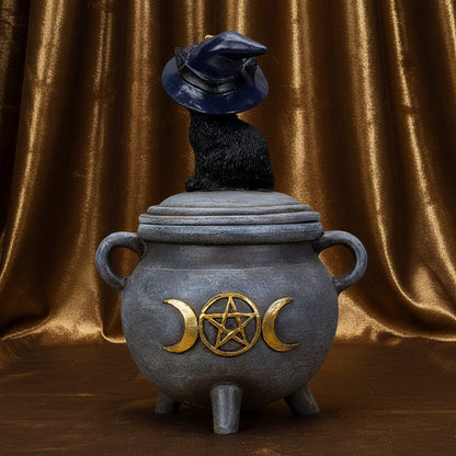 A witch-themed cauldron figurine with a black cat wearing a pointed hat sitting on top. The cauldron is dark gray with gold crescent moon and pentagram details, and has two side handles, set against a plain white background.
