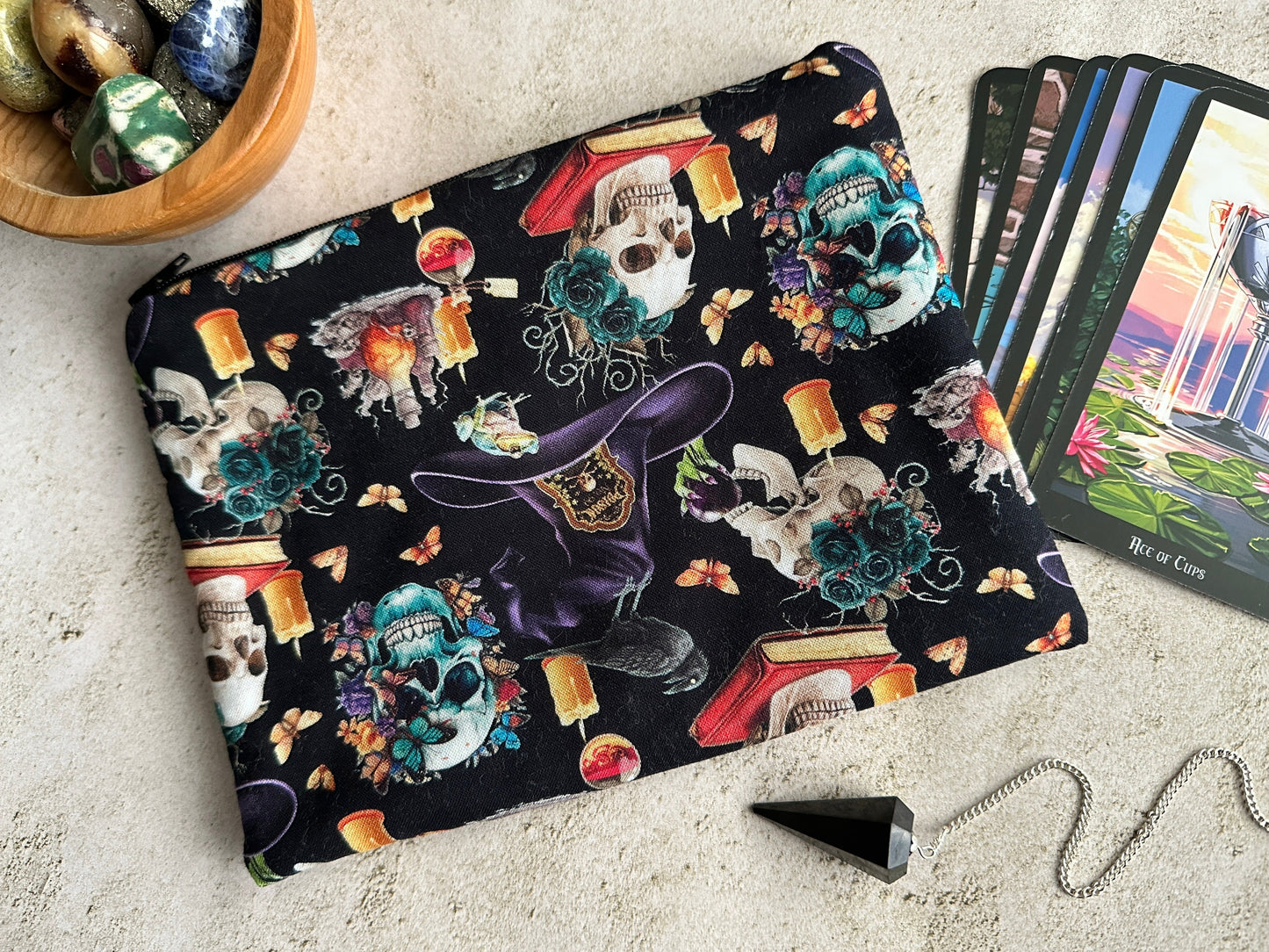 Black pouch with colorful skull and witch hat design on a light surface with tarot cards and a bowl.