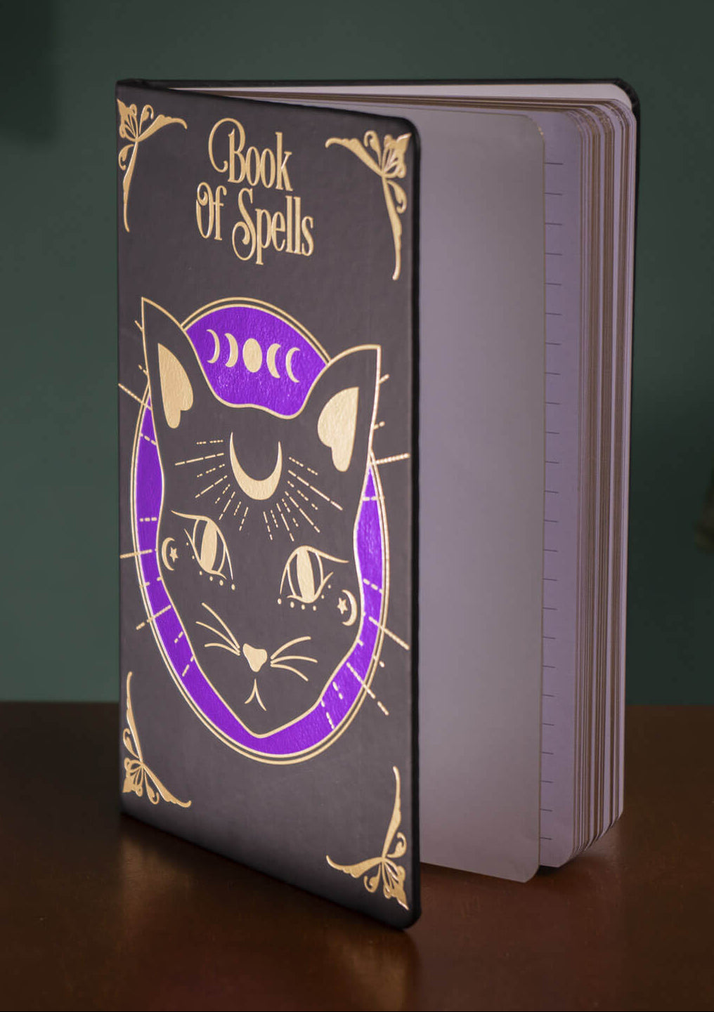 A hardcover journal titled 'Book of Spells' standing upright on a wooden table. The cover is dark gray with a black cat illustration, gold decorative flourishes, and a purple crescent moon and sun symbol.