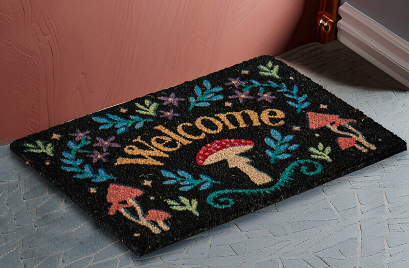 A rectangular black doormat with a colorful 'Welcome' design, featuring red and orange mushrooms, blue and green leaves, and yellow lettering, placed on a gray tiled floor in front of a pink wall.